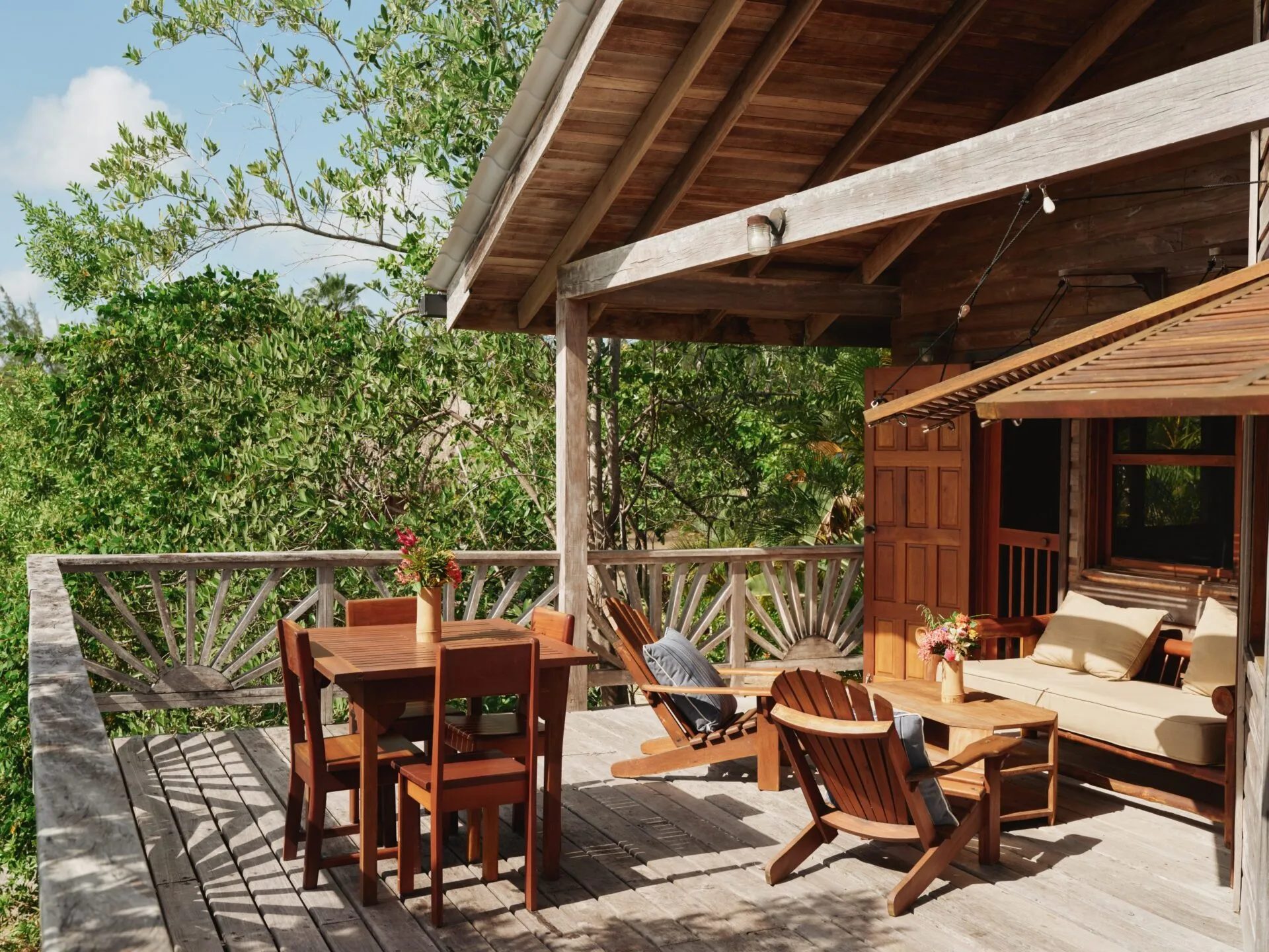 A wooden deck with Adirondack chairs, a sofa, and two tables with flowers in vases, set beneath a slanted roof surrounded by lush green trees on a sunny day.