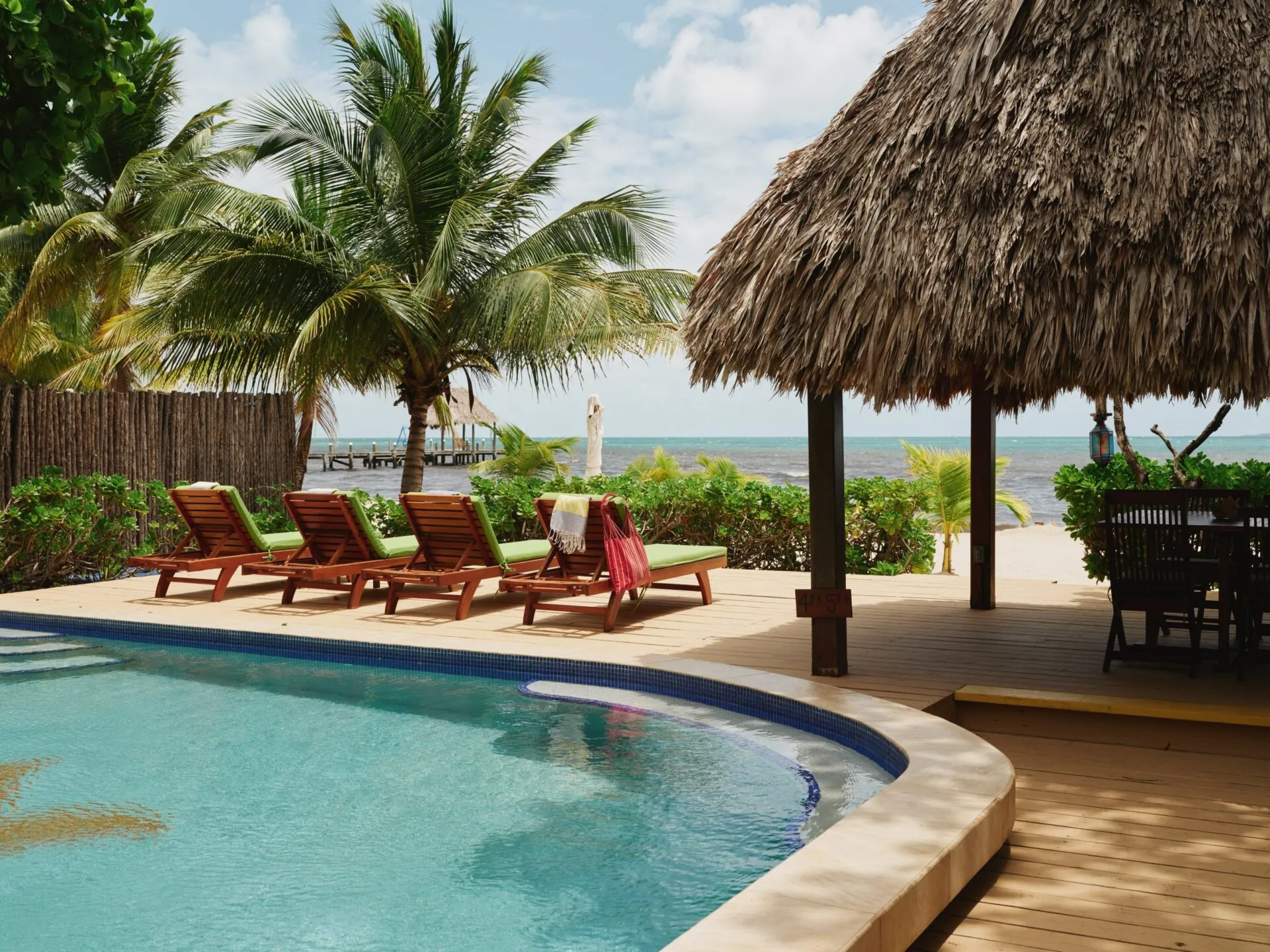 A tropical resort pool with wooden lounge chairs, palm trees, and a thatched-roof cabana, overlooking a sandy beach and the ocean at Turtle Inn, Placencia, Belize.